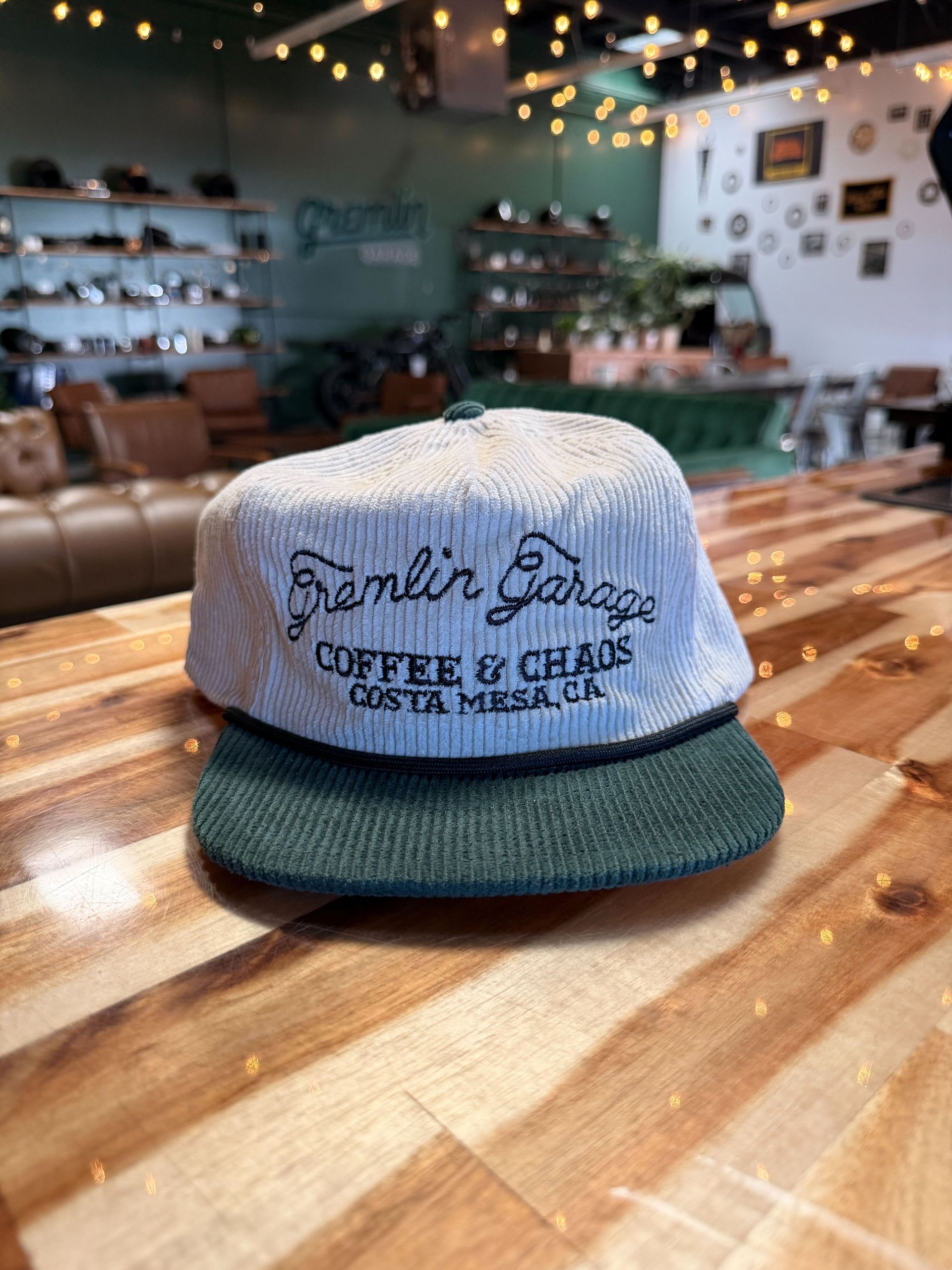 A green corduroy cap with "Gremlin Garage, Coffee & Chaos, Costa Mesa, CA" embroidered, sitting on a wooden table in a cafe.