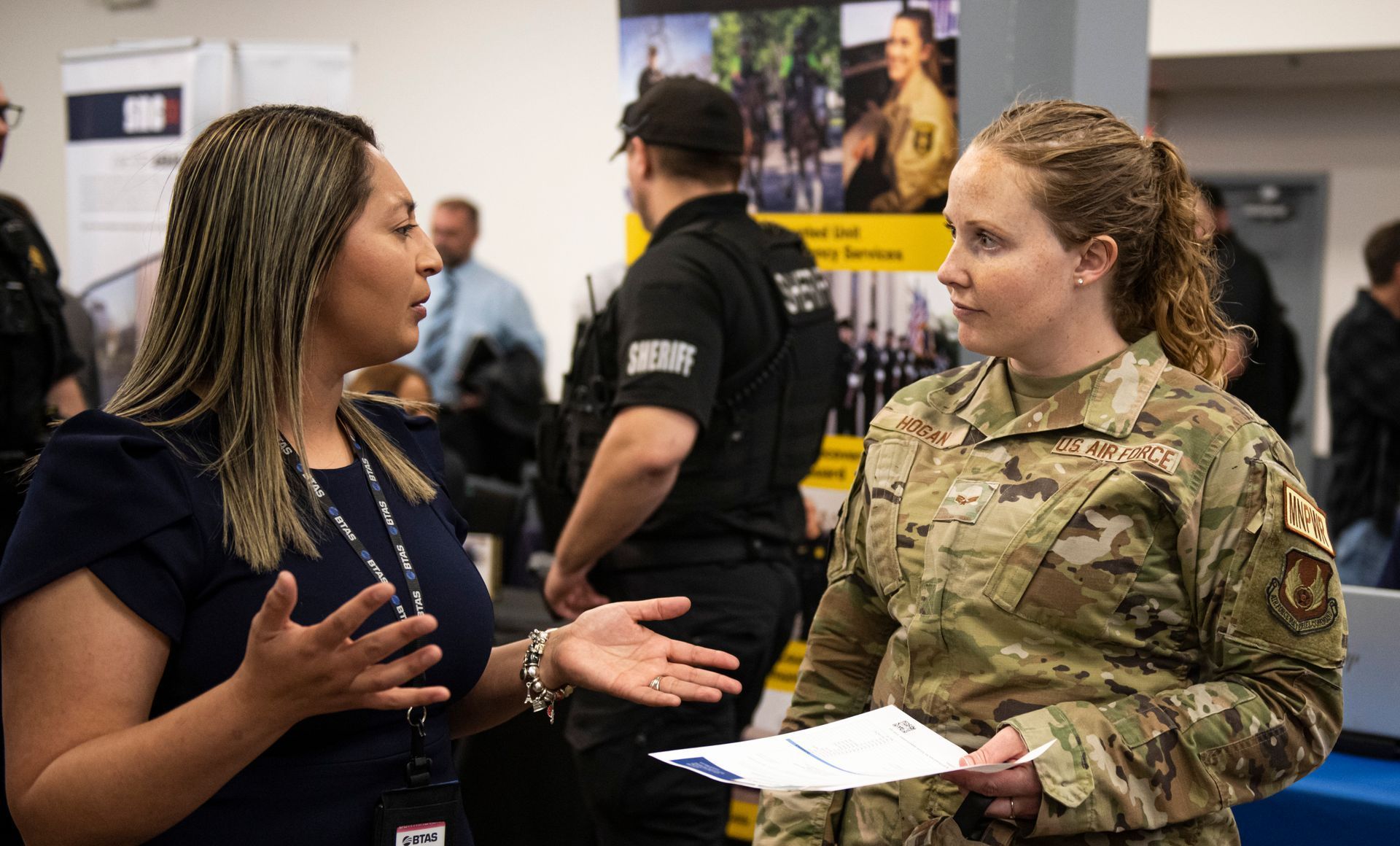 Two women in military uniforms are talking to each other at a job fair.