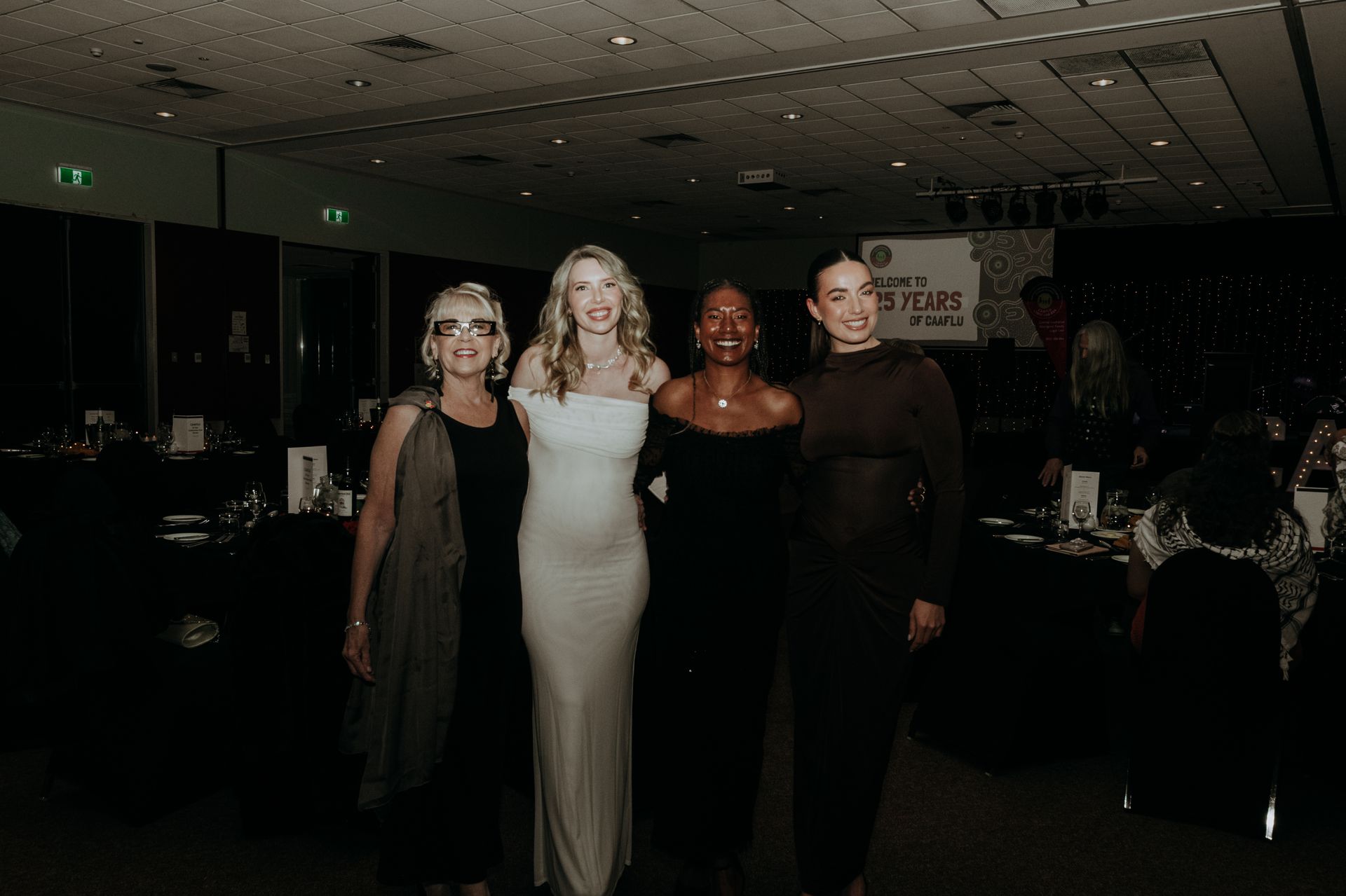 Four women pose at an event. One wears white, others in black dresses. Dimly lit CAAFLUvenue.