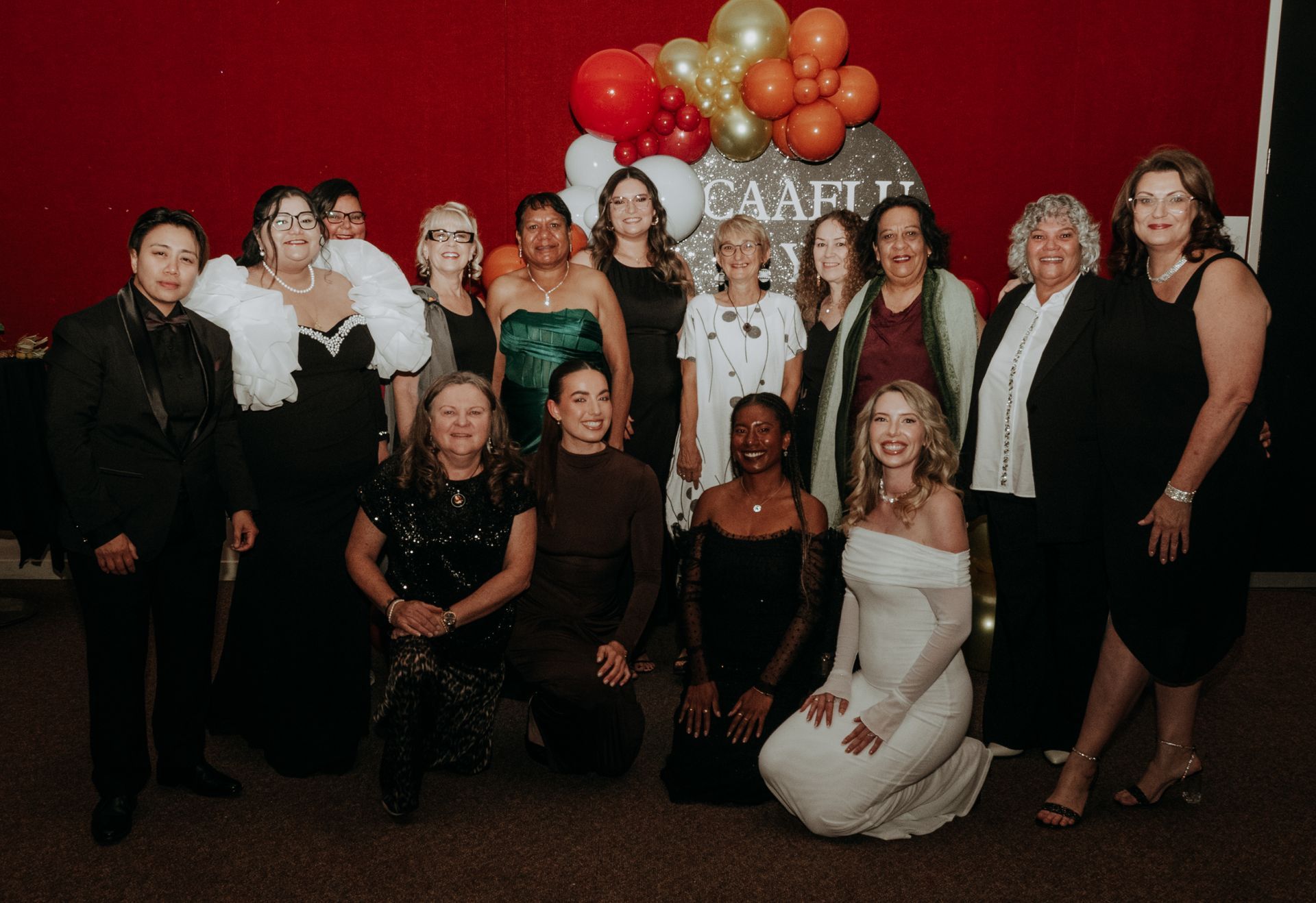 Group of people at an event, posing in front of red wall and balloons. CAAFLU