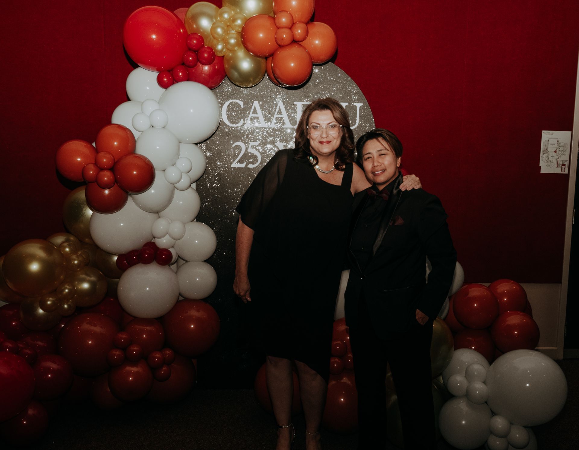 Two women smiling, posing in front of a red backdrop with balloon decorations for CAAIJ 25th anniversary.