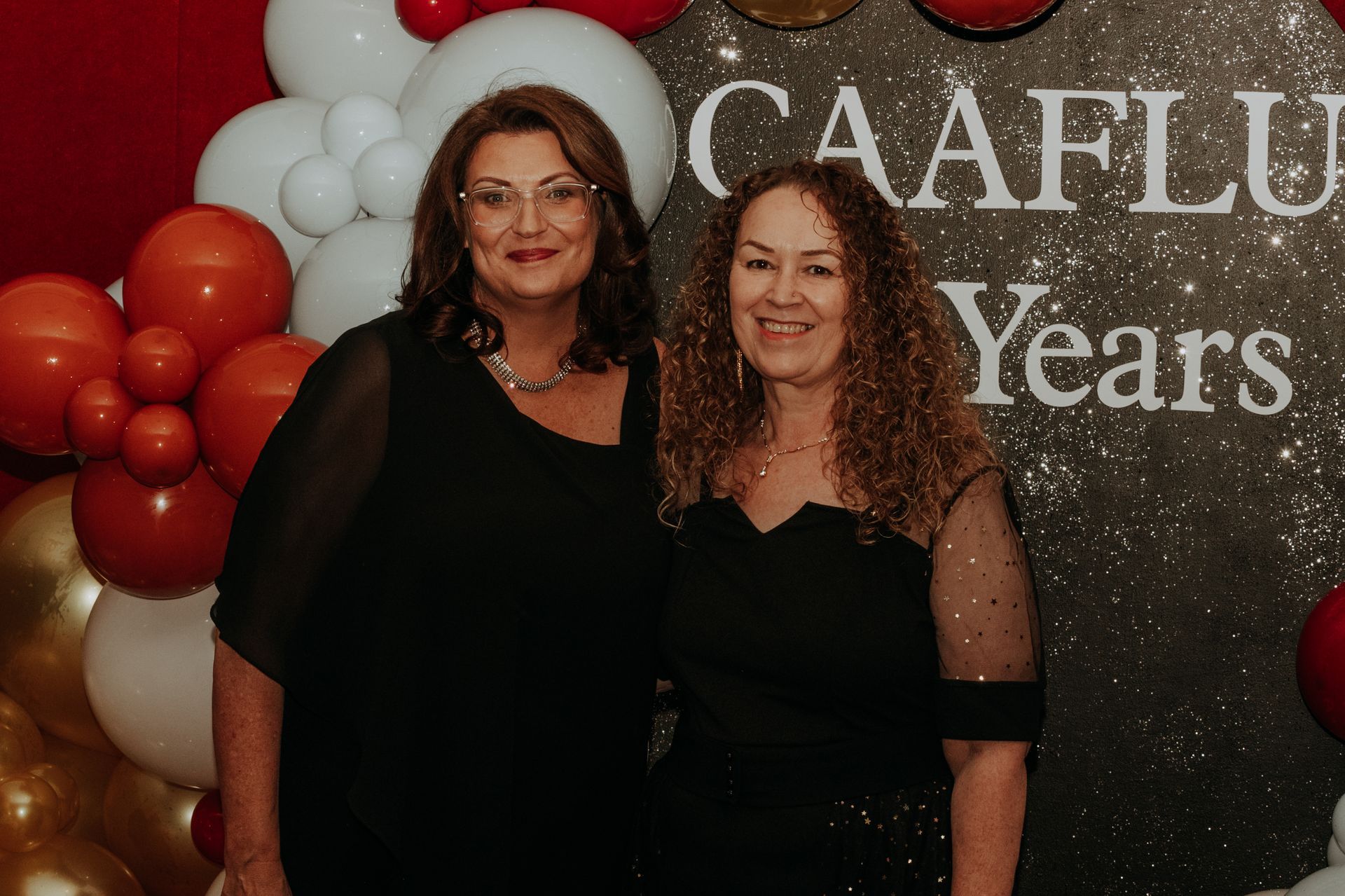 CAAFLU Two women in black dresses pose at an event with balloons and a backdrop that reads 