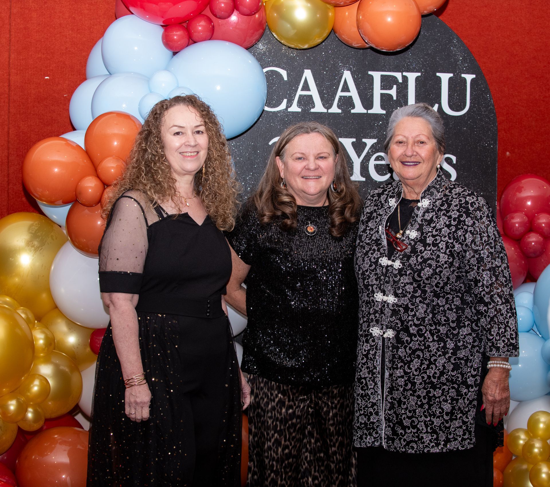 Three women at a party with balloons. Backdrop says 