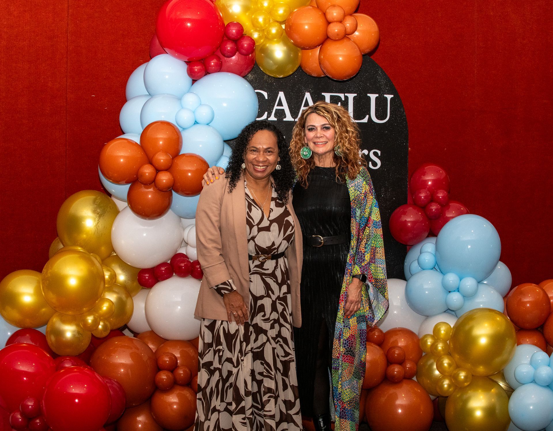 Two women smiling, posing by colorful balloons at an event. CAAFLU