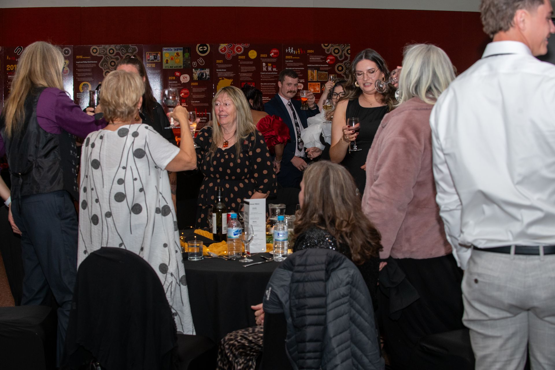 People at an event, gathered around tables with drinks and snacks, inside a room with red walls. CAAFLU