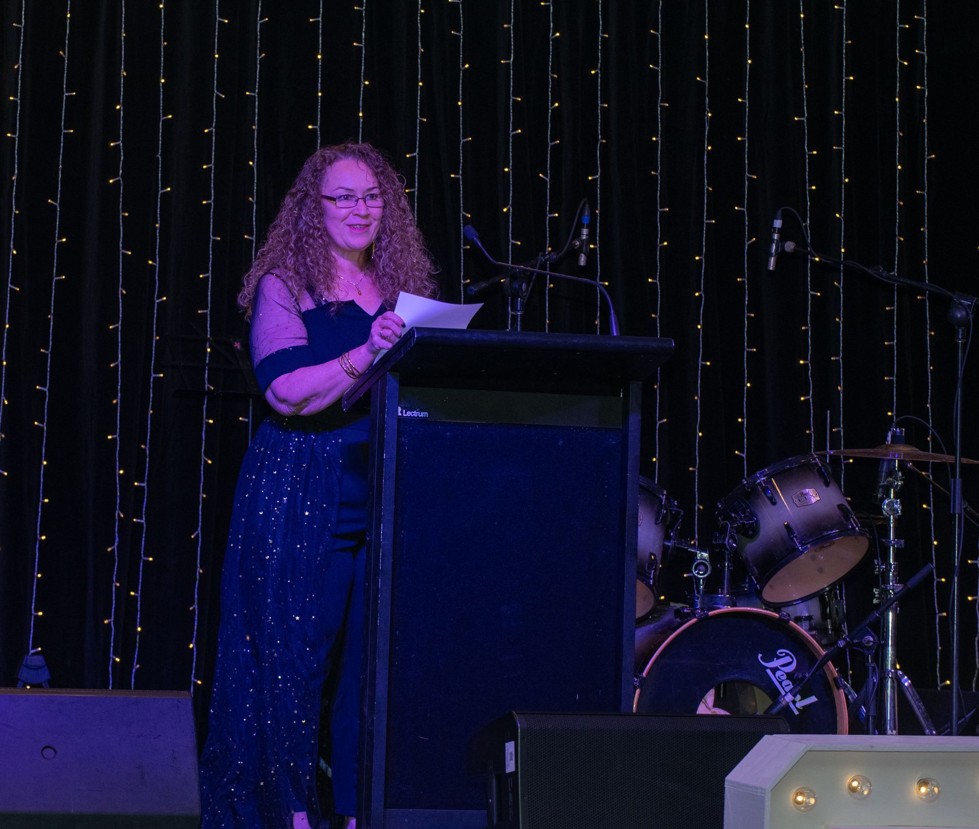 CAAFLU Woman with curly hair in a formal gown speaking at a podium, lit stage with string lights and drum set.