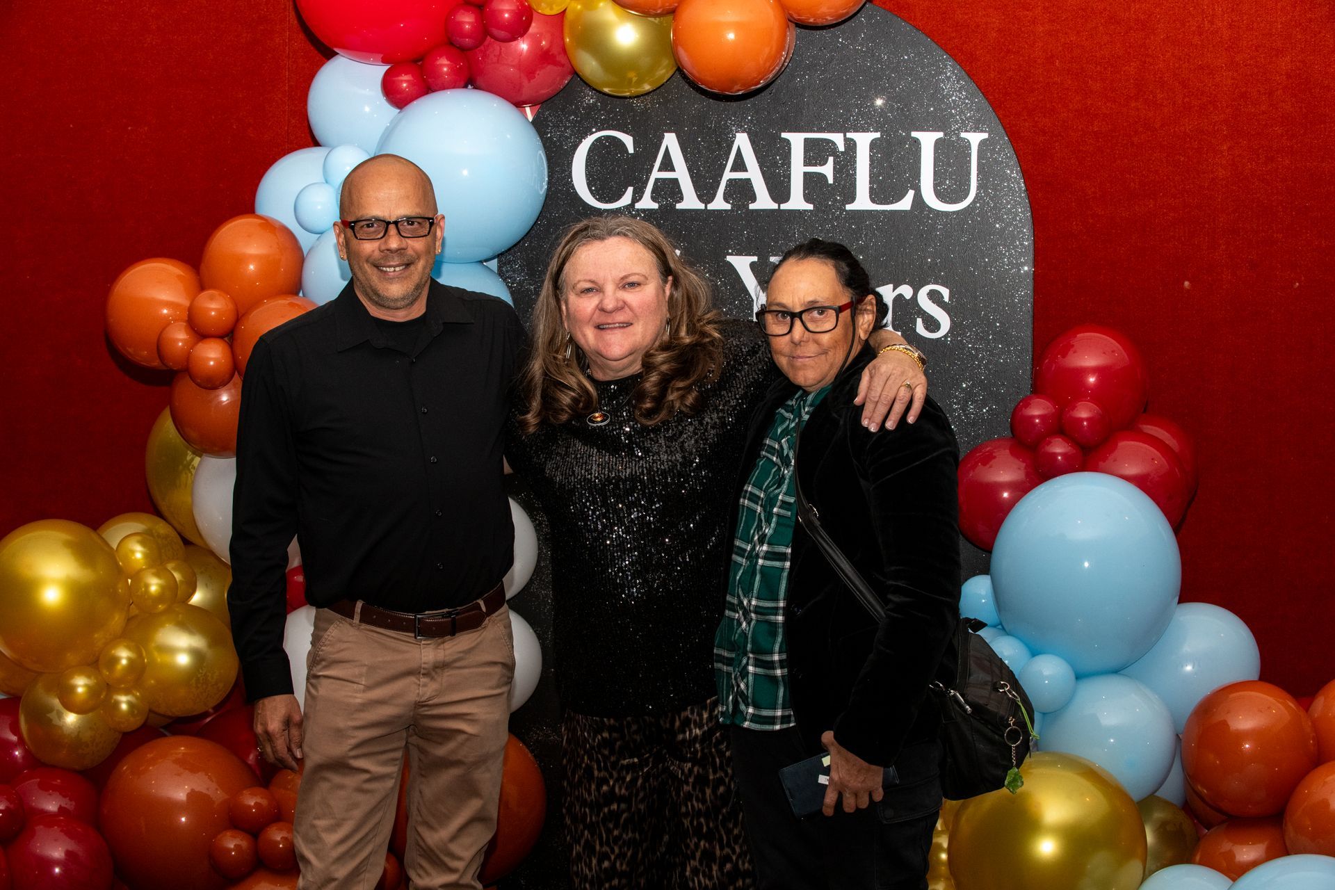 Three people smiling, posing in front of a red backdrop with balloons. The word 