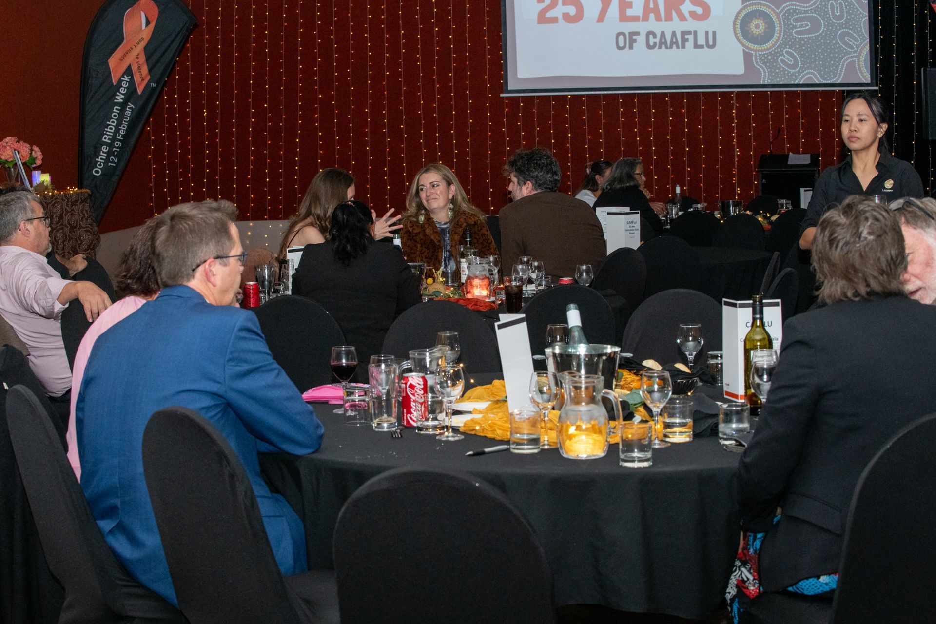 People at a formal event seated at round tables, with a server, a backdrop CAAFLU
