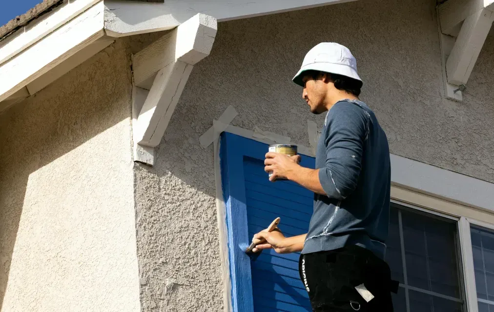 Man in hard hat paints blue shutter on a house.