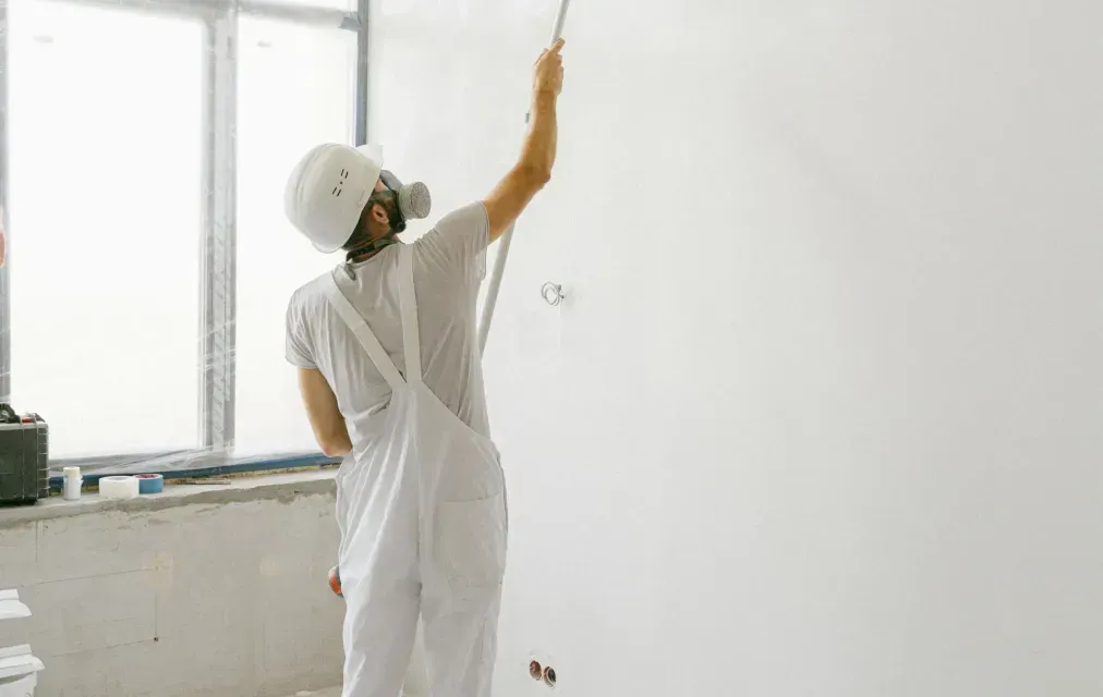 Person in white protective suit spraying paint on a white wall in a room.