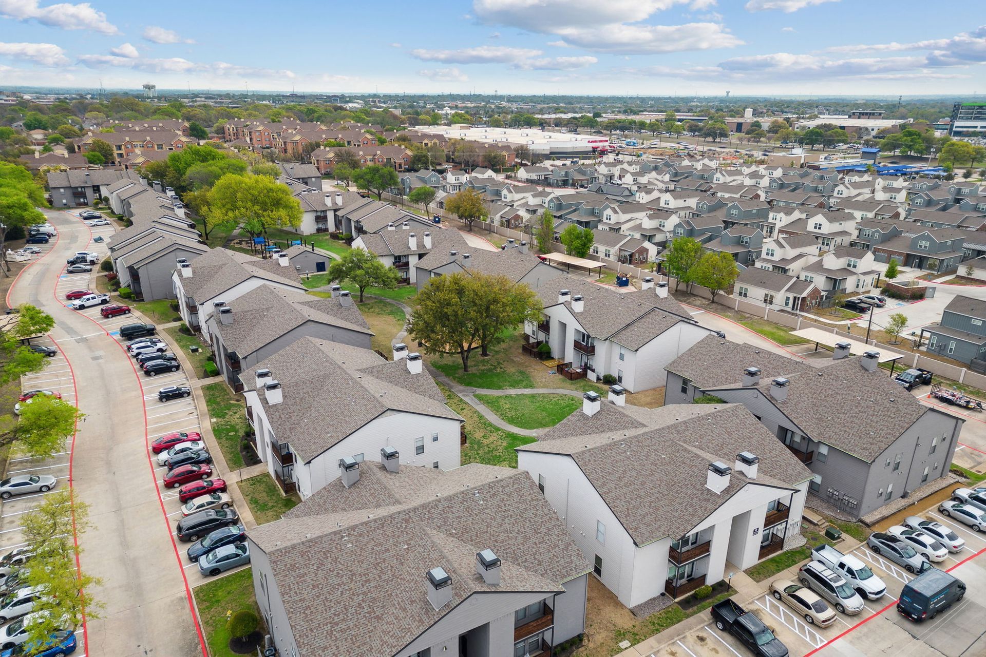 Aerial view of a suburban apartment complex with gray roofs, parked cars, and a tree-lined courtyard.