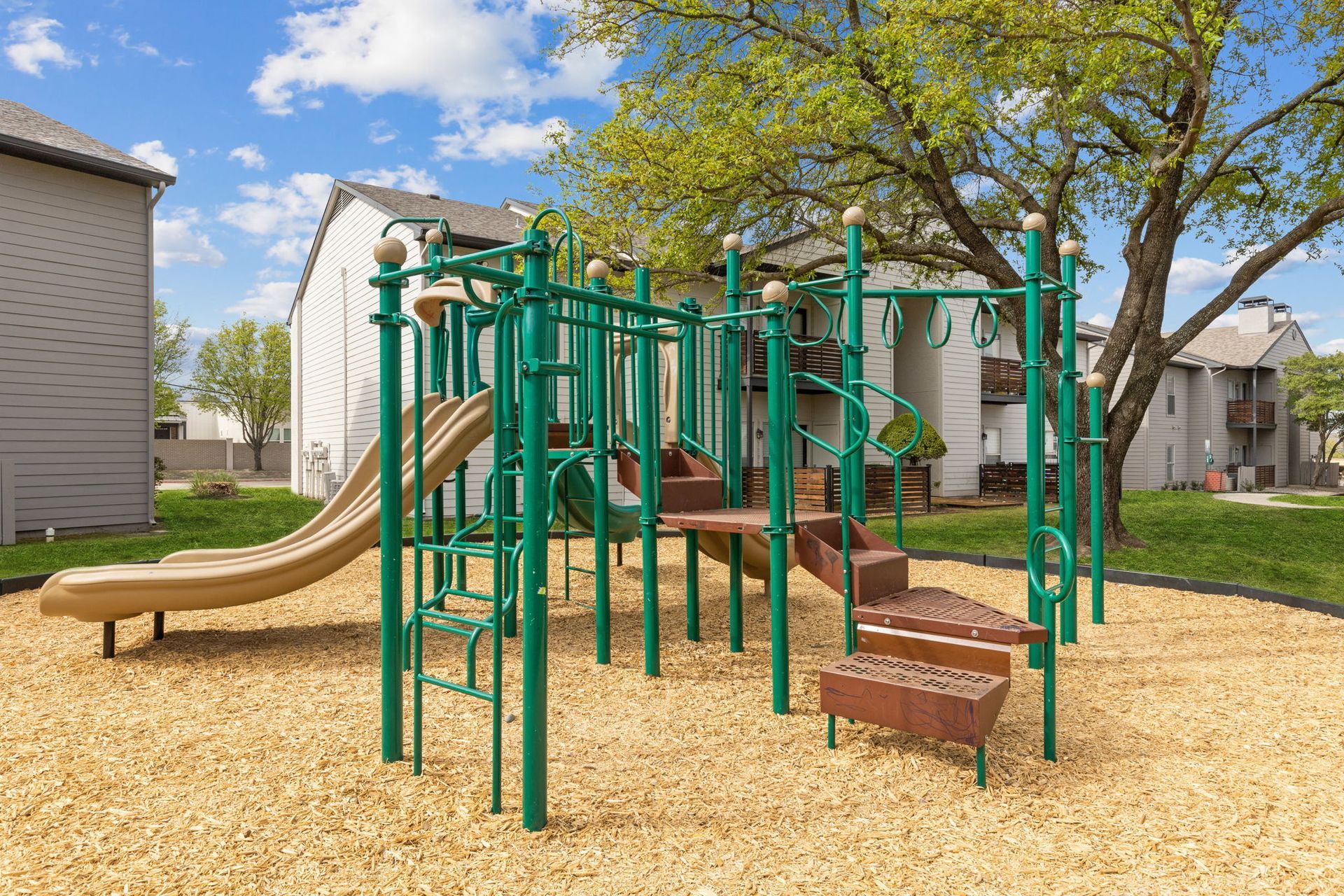 Green playground with a slide and stairs on wood chips, by an apartment building.
