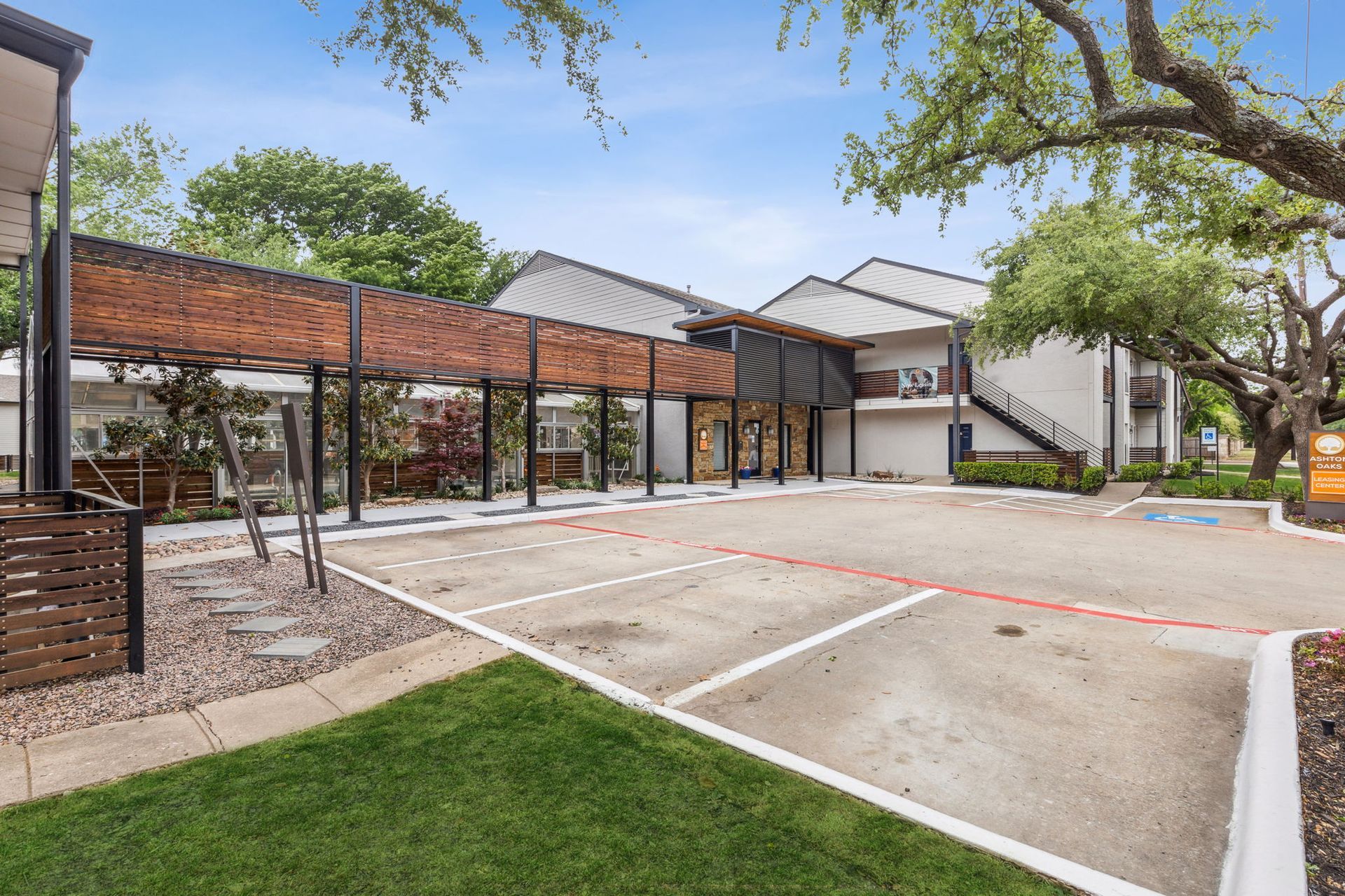 Apartment building exterior with parking, covered walkway, and green lawn under a blue sky.
