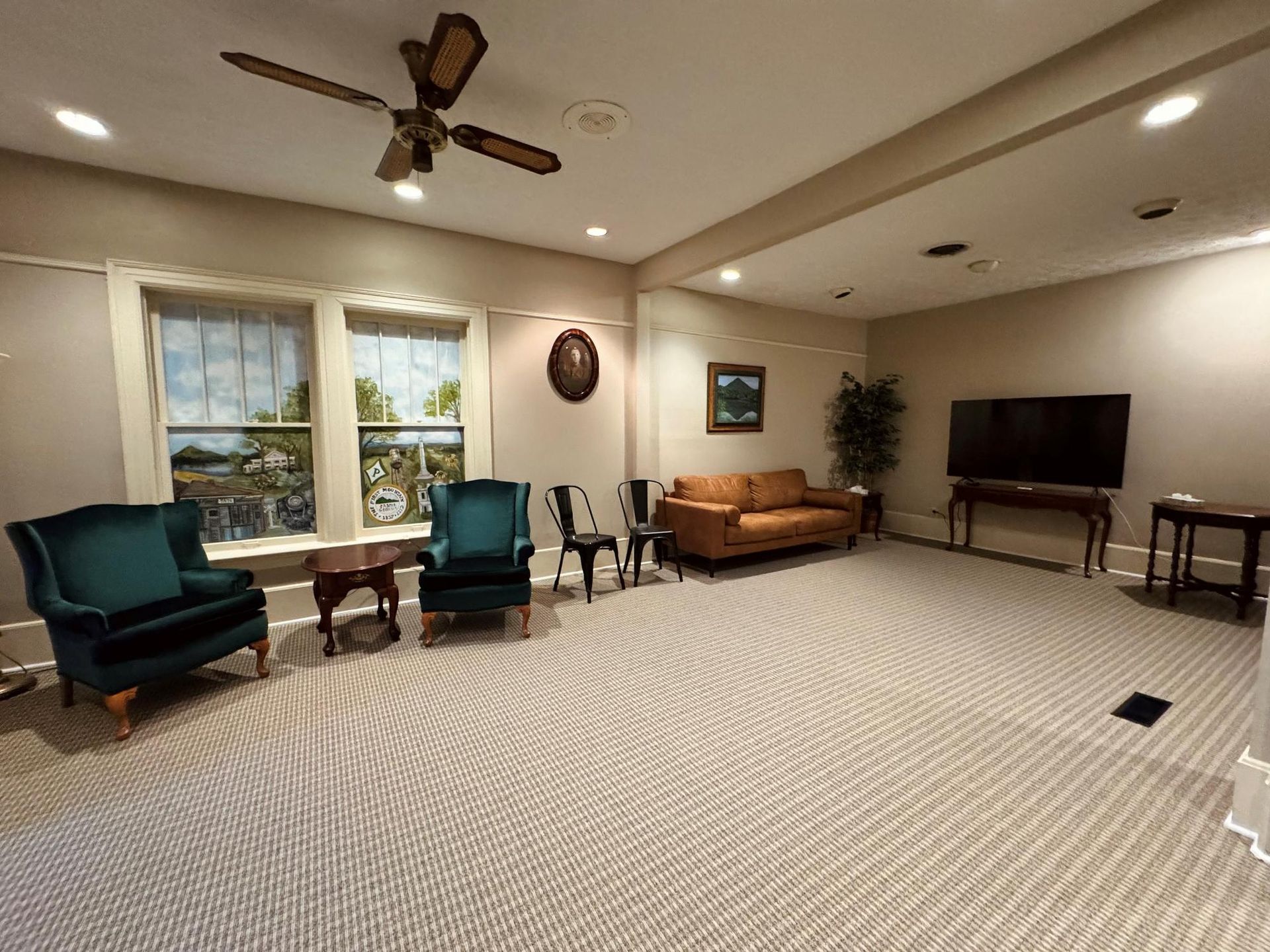 Bright lounge with teal chairs, tan sofa, round tables, TV, and a large window on patterned carpet.