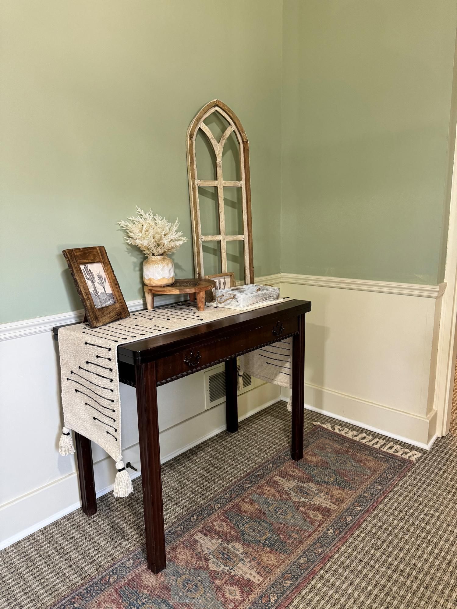 Small entryway table with mirror, framed photo, flowers, and decorative tray against sage green walls.