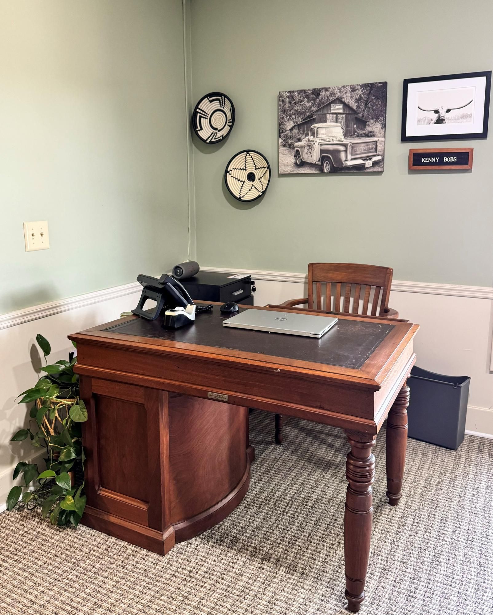 Home office with wooden desk, black phone, laptop, wall art, and potted plant against a light green wall