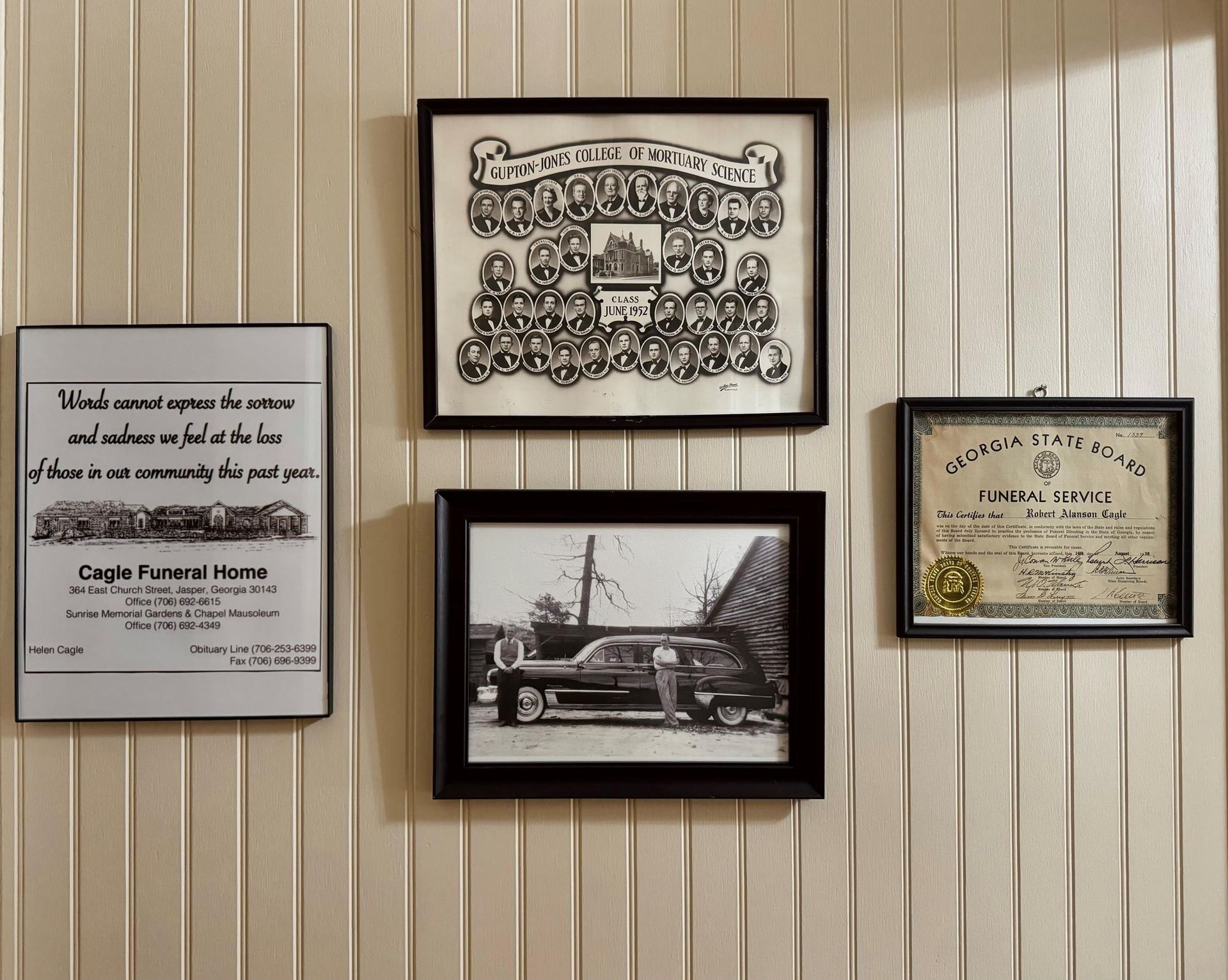 Four framed certificates and a black-and-white car photo mounted on a beige paneled wall.