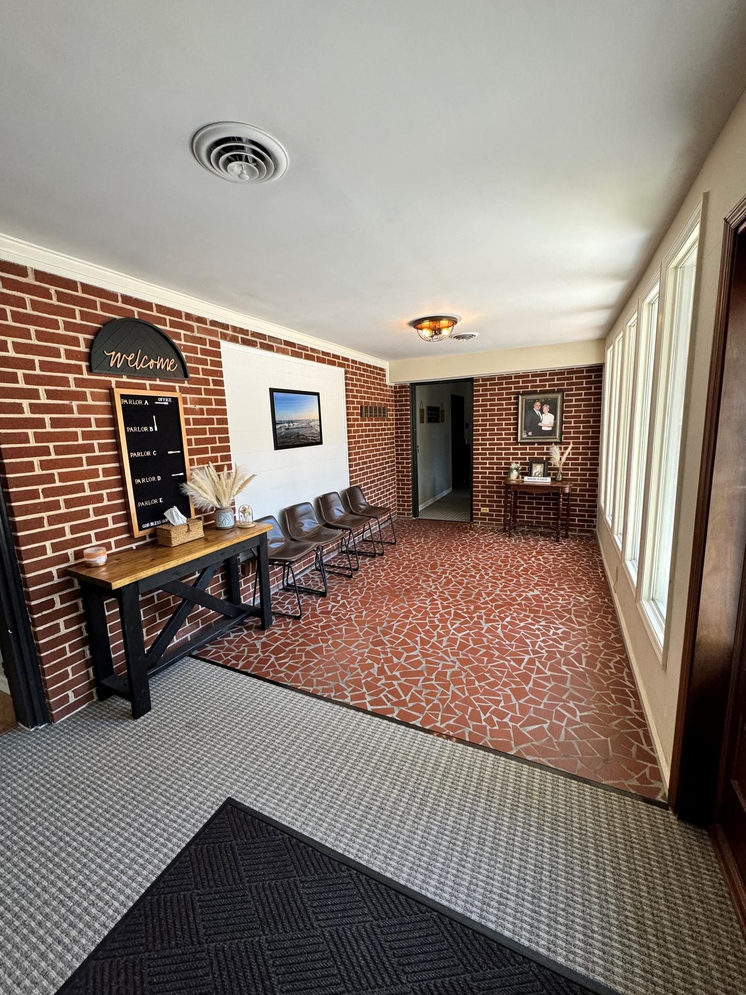 Brick-walled waiting area with chairs, a table, windows, and patterned carpet in a hallway