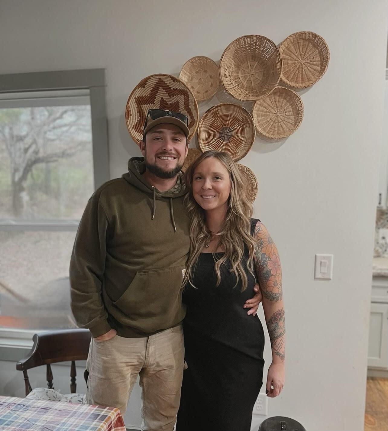 Two people smiling indoors beside a wall of woven basket decor