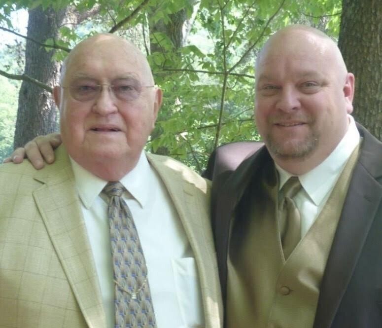 Two men in suits smiling outdoors, with trees in the background.
