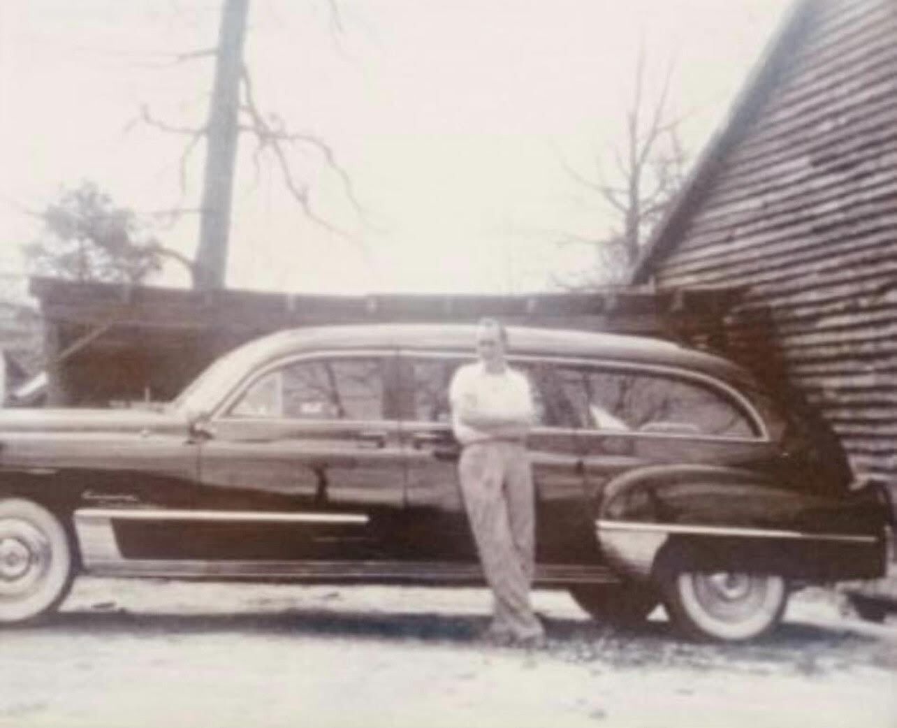 Vintage black station wagon parked beside a house with a man leaning against it.