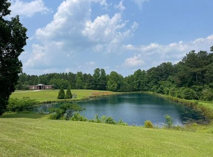 Grassy hill overlooking a small blue pond beside a wooded field under a partly cloudy sky
