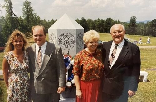 Four adults posing outdoors in formal attire beside a memorial monument on a grassy lawn.