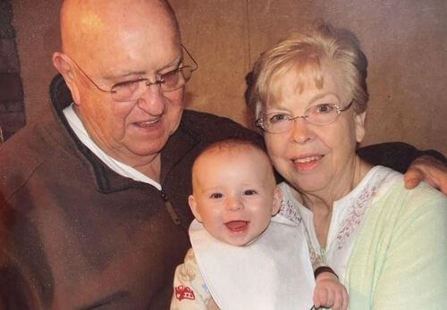 Smiling baby held between an elderly man and woman, all seated close together indoors.