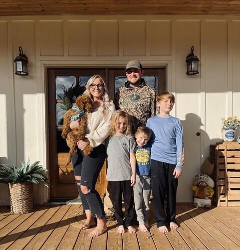 Family of five standing on a sunlit porch, smiling in front of a front door.