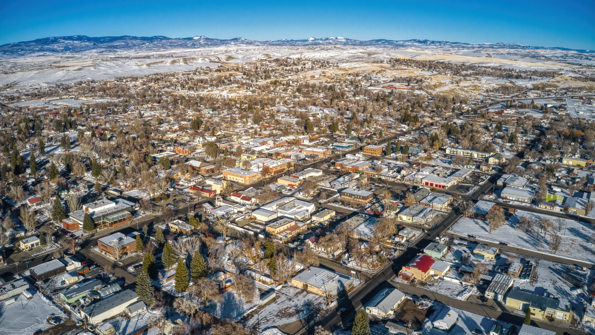 Snow-covered suburban neighborhood spreading toward distant mountains under a clear blue sky.