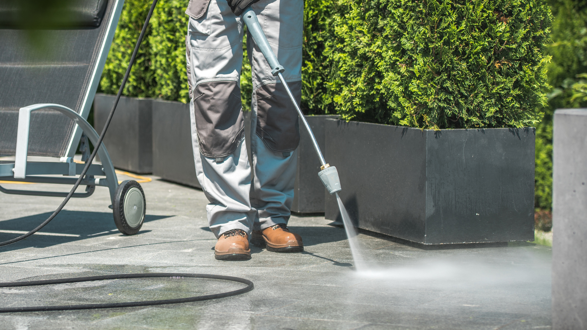 Worker pressure washing pavement beside a hedge, wearing gray pants and brown boots.