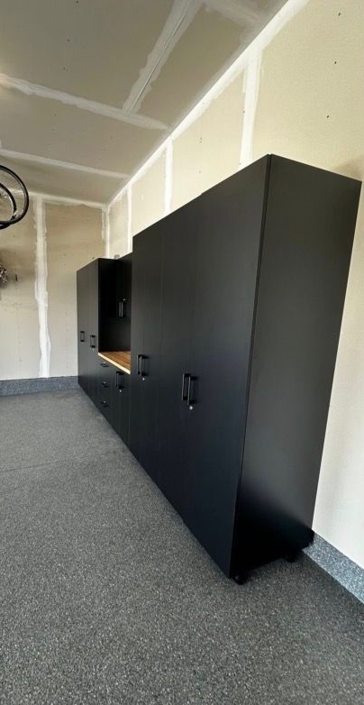 Black storage cabinets along a garage wall in a carpeted room with unfinished walls and ceiling