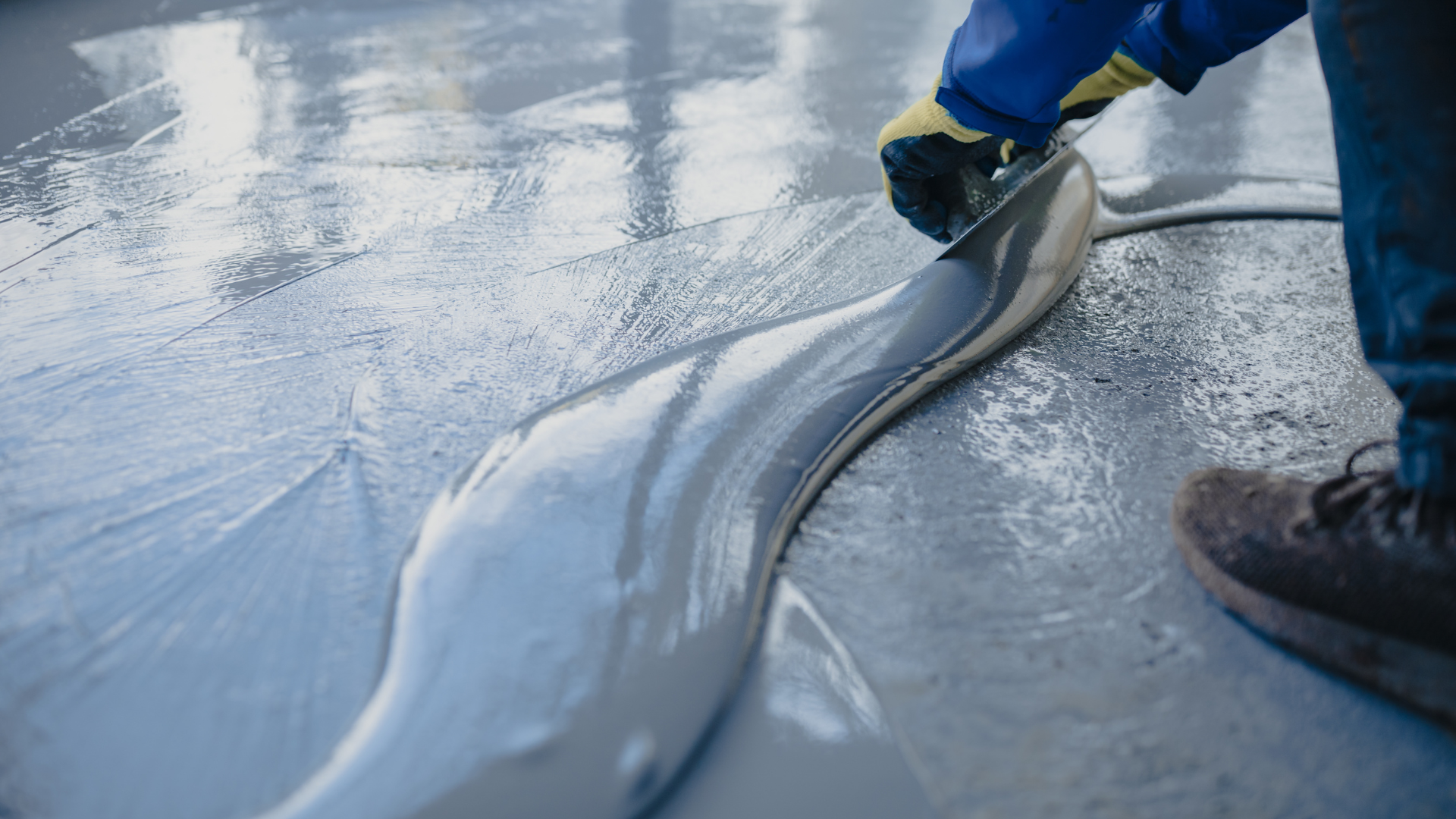 A worker wearing gloves spreads wet gray epoxy resin across a floor using a manual squeegee tool.