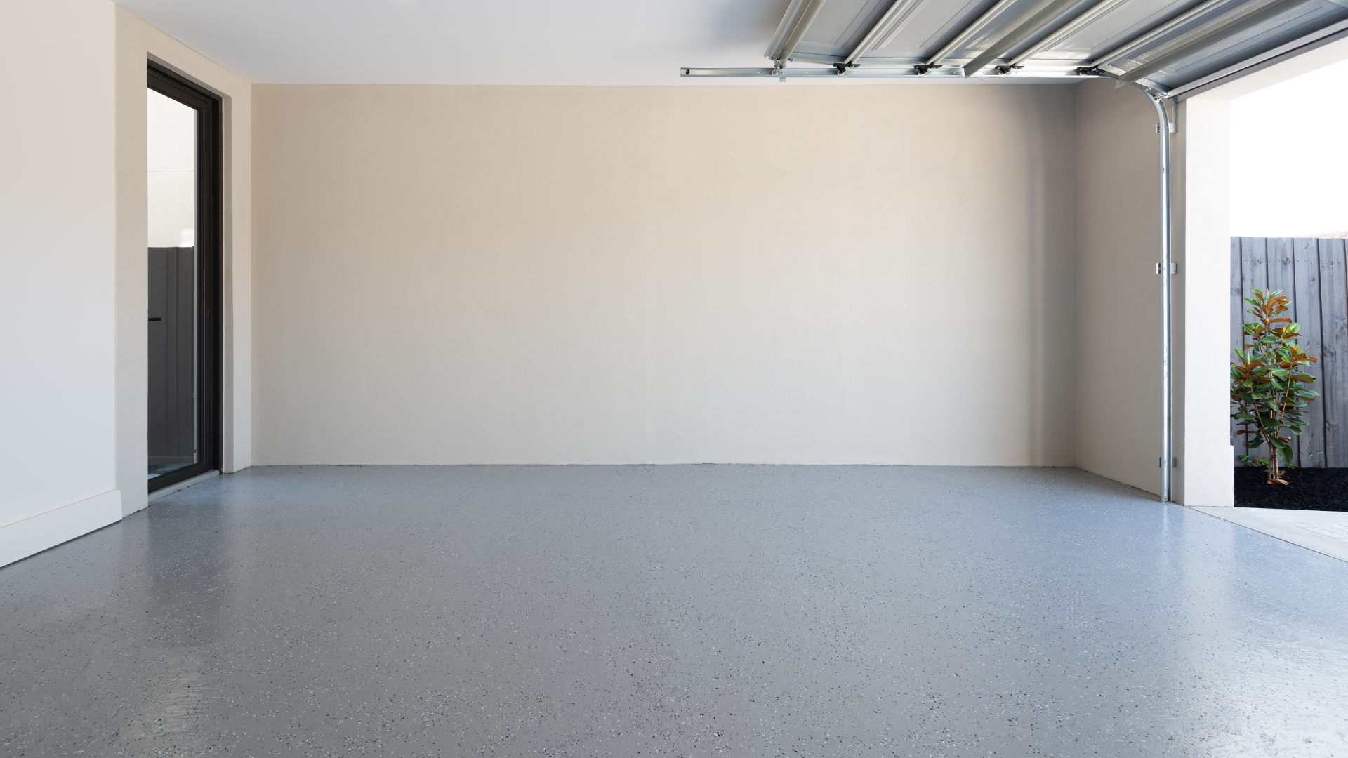 Empty modern garage with white walls, speckled floor, and open doorway showing daylight and plants