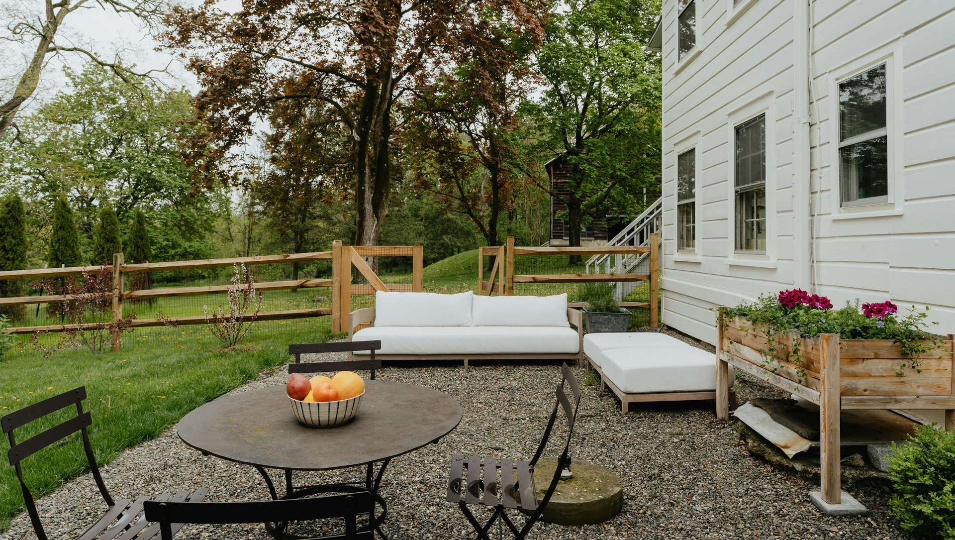 Backyard patio with white house, wicker chairs, fire pit, and cushioned bench by a wooden fence