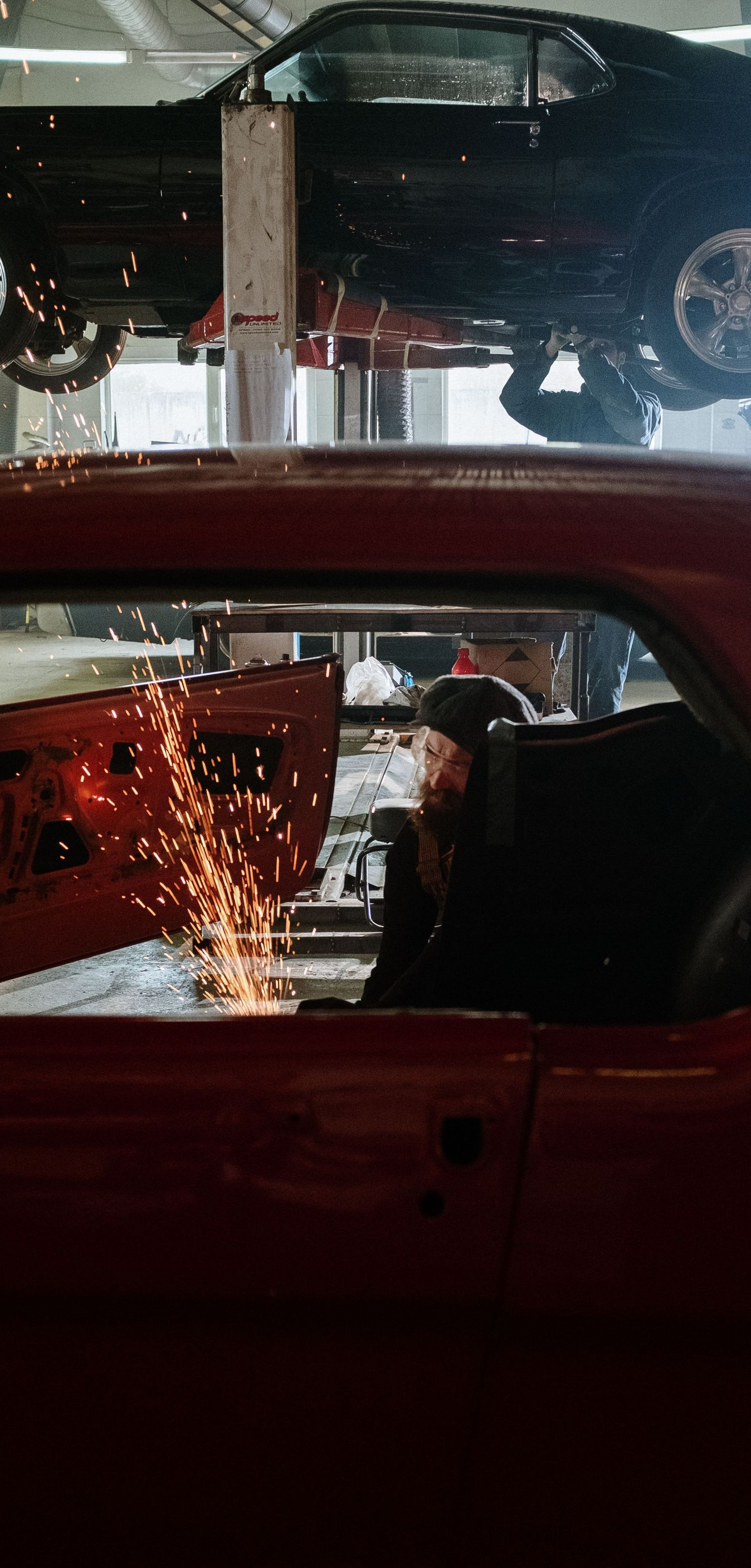 Cars in a repair shop, one lifted on a hoist above a red car with sparks nearby.