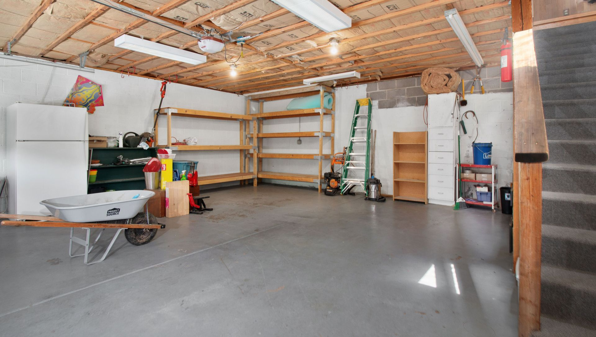 Spacious garage with concrete floor, shelving, tools, and a white freezer under exposed wooden ceiling.
