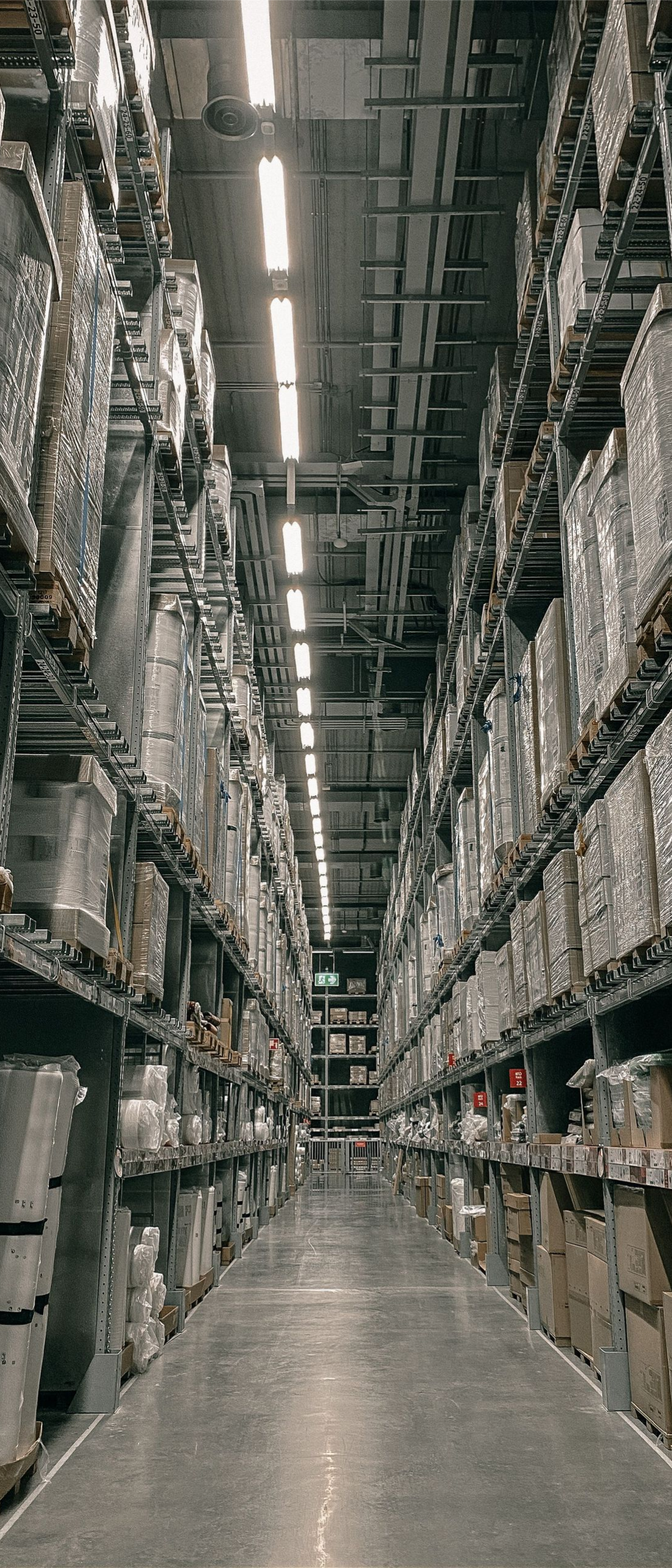 Aisle in a warehouse with tall stacked shelves of boxes and goods under bright overhead lights.