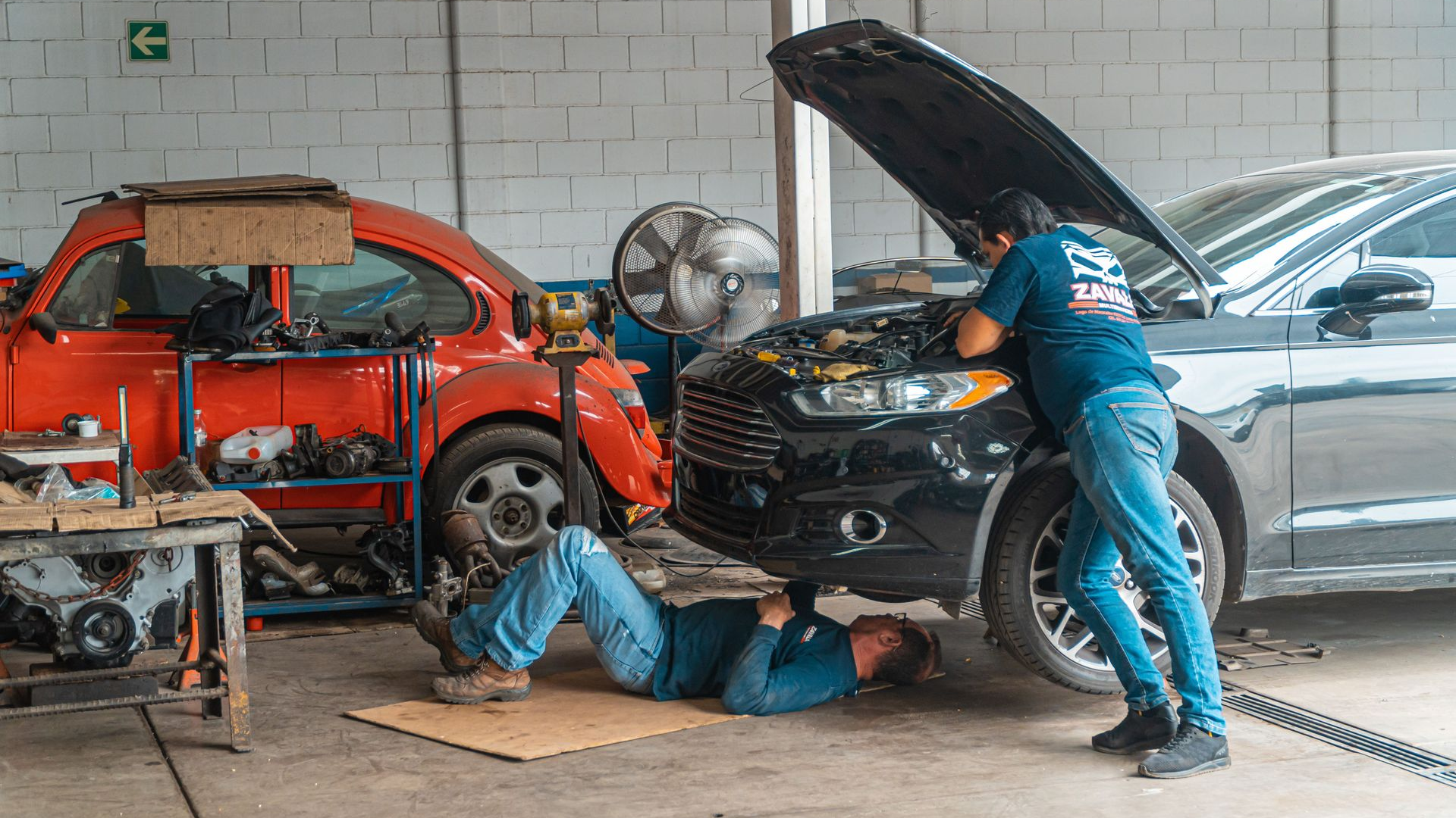 Mechanic under a car in a garage while another works with the hood open; red car and fan nearby
