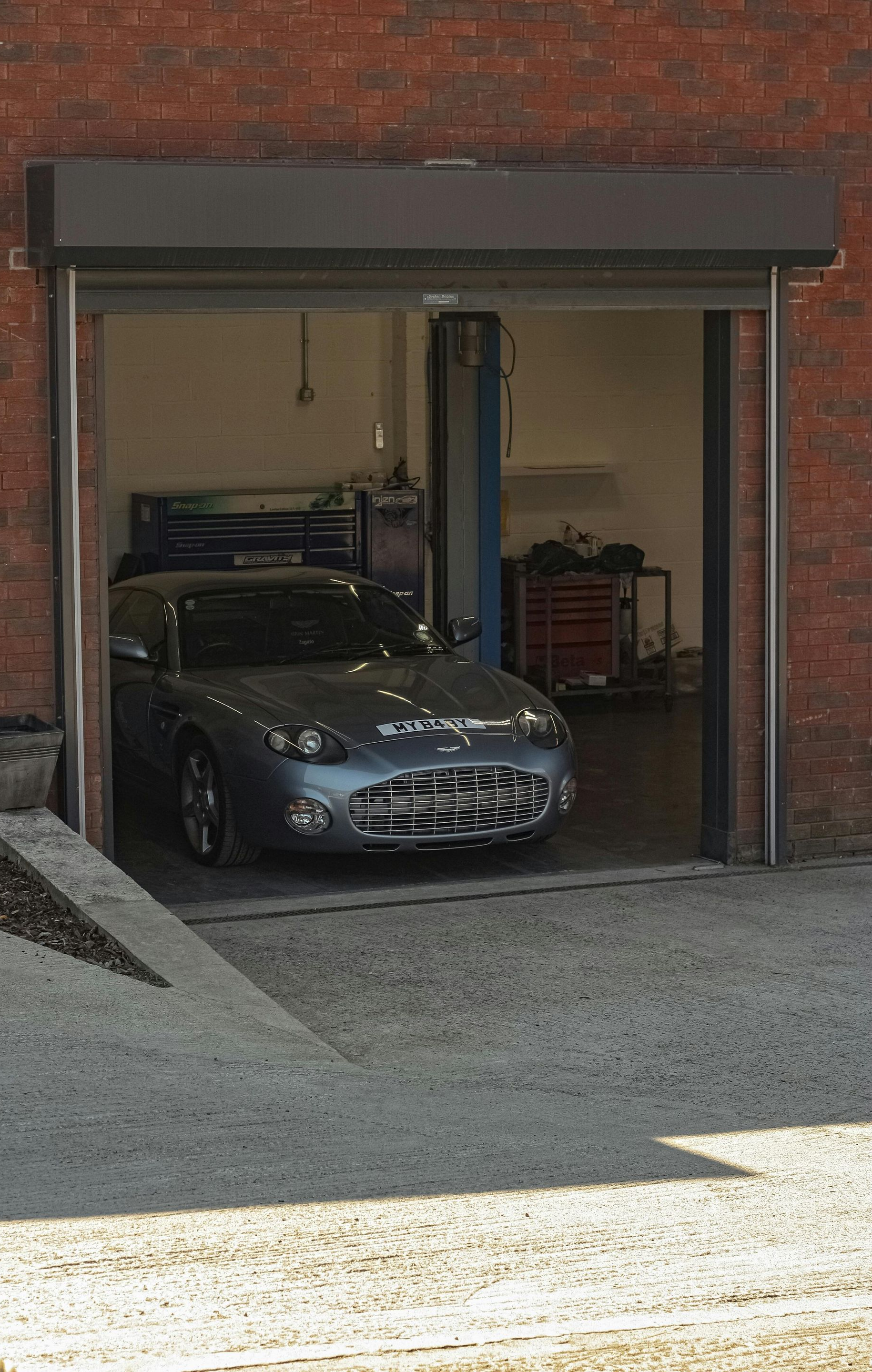 Gray sports car inside a garage with the door open, viewed from outside on a gravel driveway