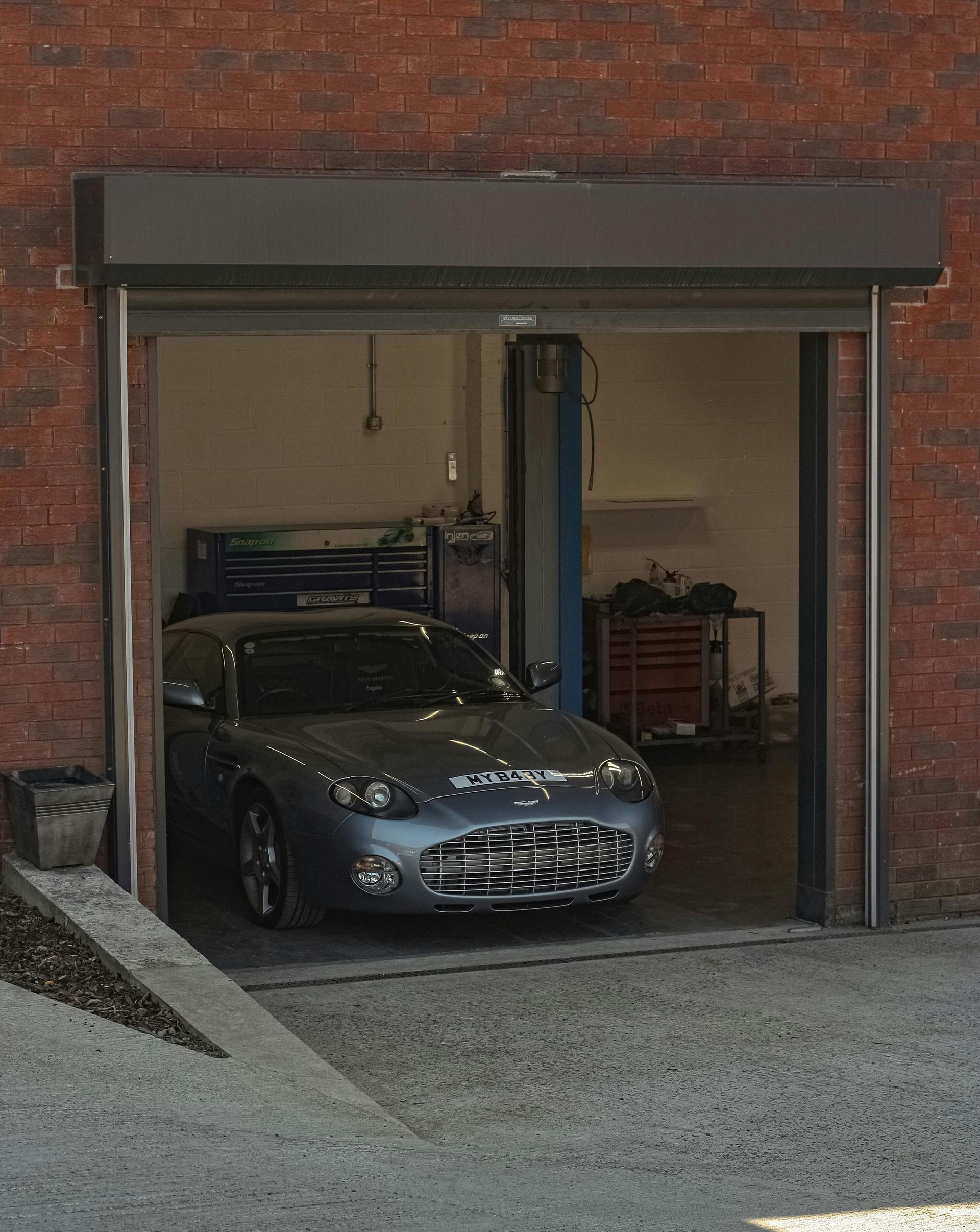 Silver sports car parked in a partially open garage with a brick exterior.