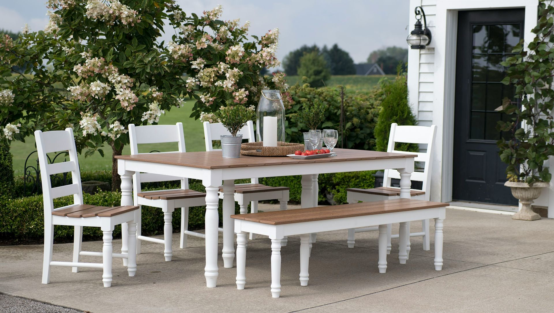 White outdoor dining set on a patio beside a house, with a table, benches, and flowering shrubs.