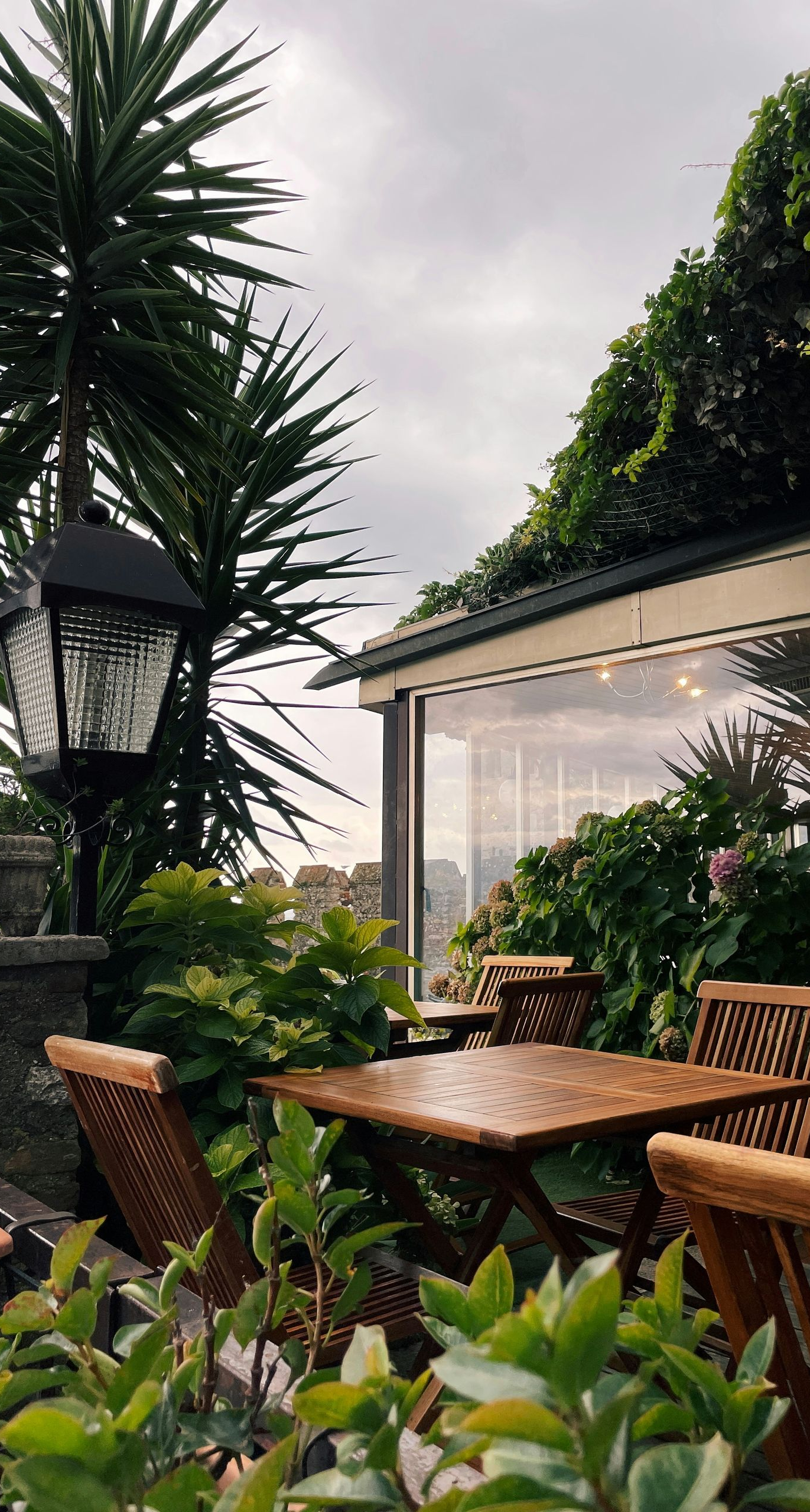 Cozy outdoor patio with wicker chairs, a wooden table, and lush greenery beside a glass-walled house at dusk