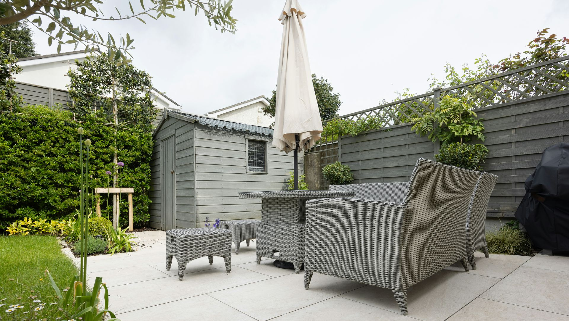 Modern backyard patio with wicker seating, umbrella, and gray shed beside green lawn and hedges