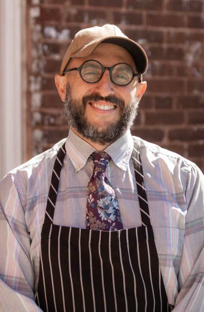 Aaron Rietz in glasses, cap, and striped apron standing against a brick wall