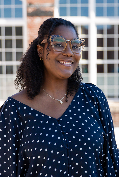 Brittney Adams smiling outdoors in a navy polka dot blouse and glasses, standing before a white building.