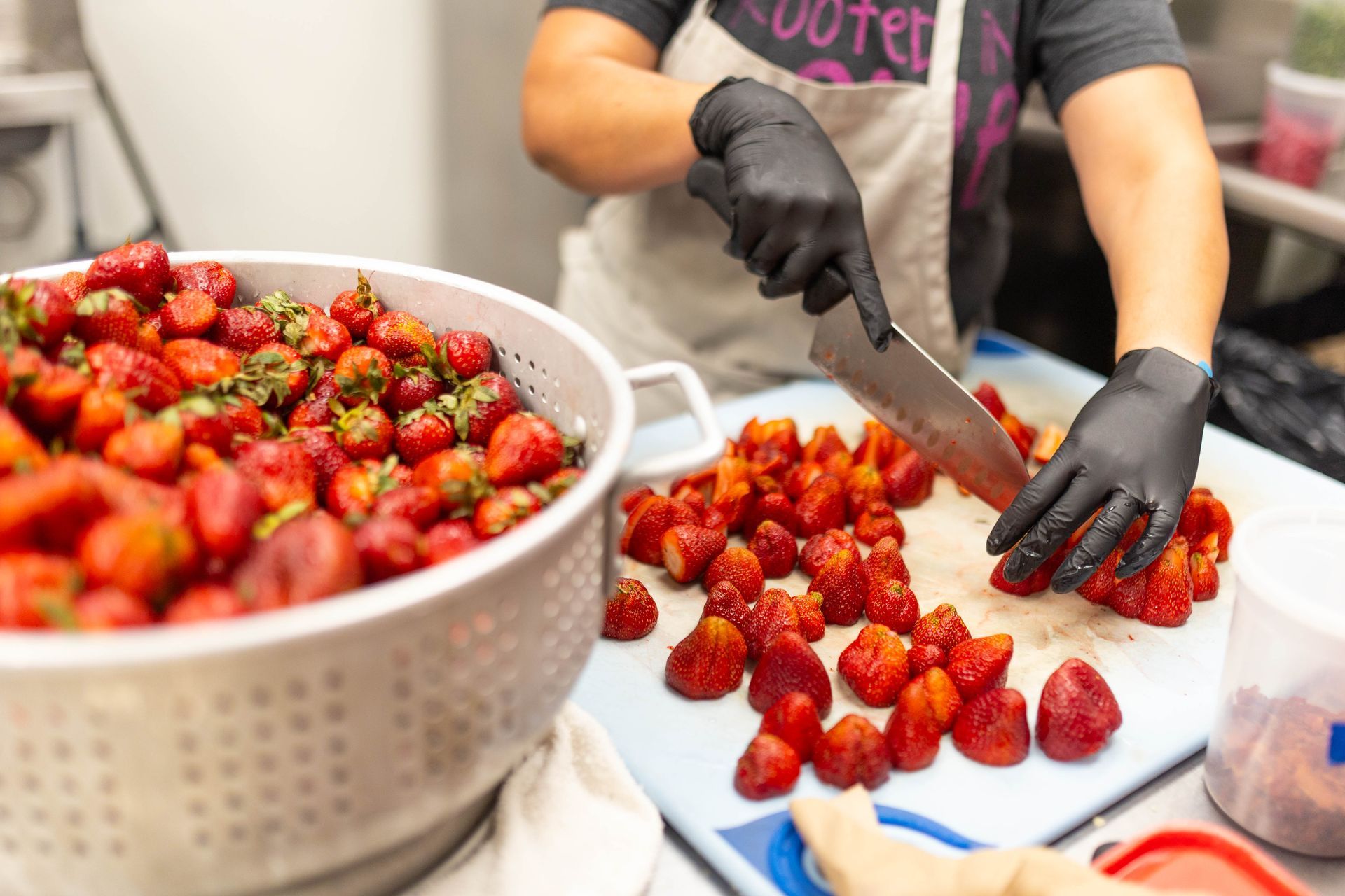 Gloved worker slicing strawberries on a tray beside a large bowl of berries.