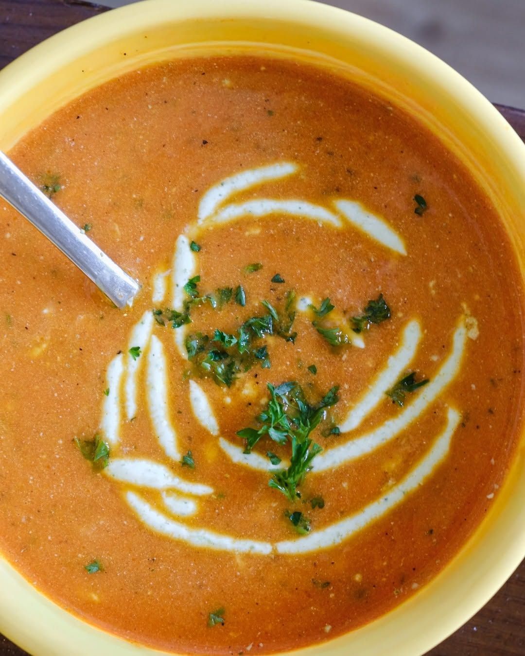 Bowl of orange soup topped with white cream swirls and chopped herbs, with a spoon at the side
