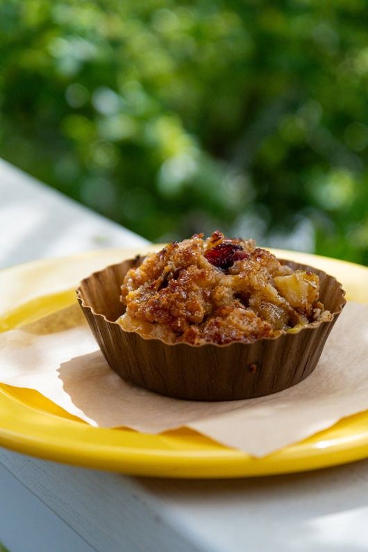 A crumb-topped muffin in a brown liner on a yellow plate outdoors with blurred green background