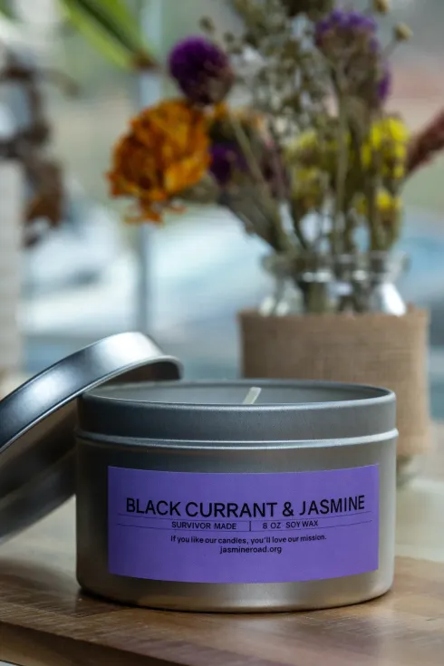 Black currant and jasmine candle in a gray tin on a wooden table with dried flowers behind it